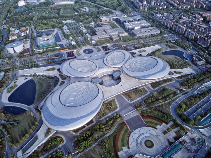 An aerial view of a modern, circular, and white building complex surrounded by green spaces and a body of water, with a cityscape visible in the background.