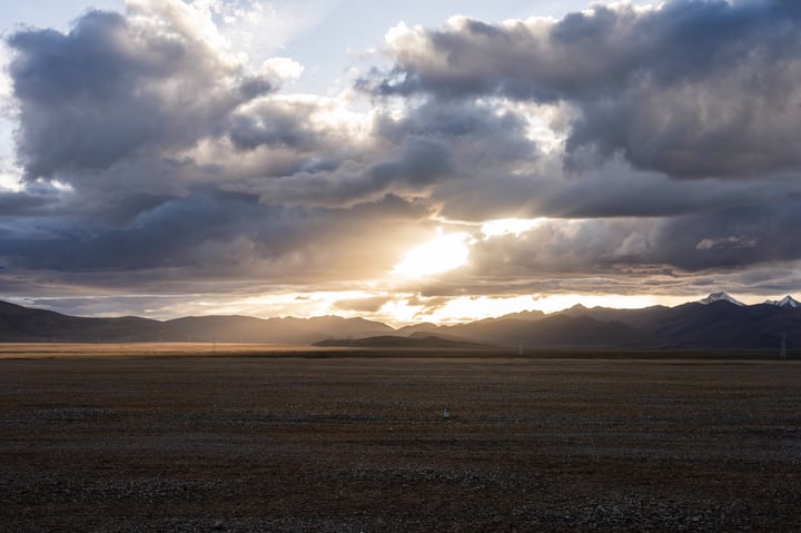 A vast, barren landscape stretches out under a dramatic sky, with a solitary light source casting a warm glow on the horizon. The sun is partially hidden behind dark clouds, casting a dramatic shadow on the landscape. The mountains in the distance are partially obscured by the clouds, adding depth to the scene.