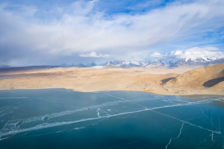 A serene landscape features a deep blue lake reflecting the sky, with snow-capped mountains in the distance. The sky is filled with white clouds, and the landscape is dominated by brown and beige tones.