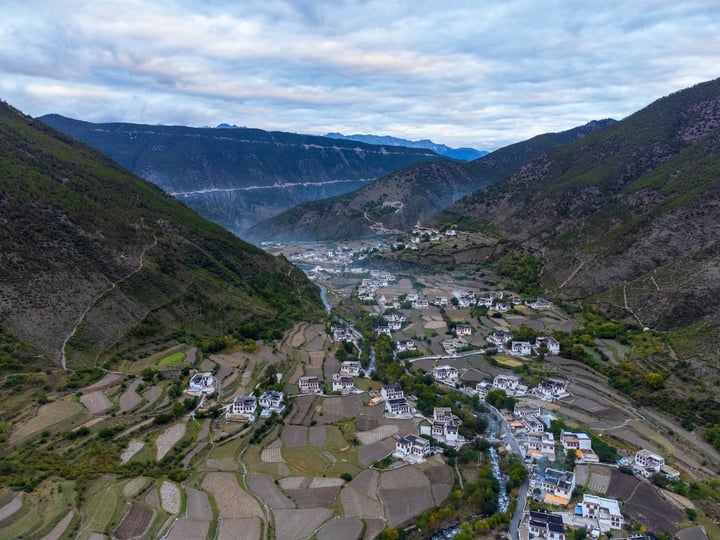 An aerial view of a valley with a small village, surrounded by mountains and greenery, with a cloudy sky above.