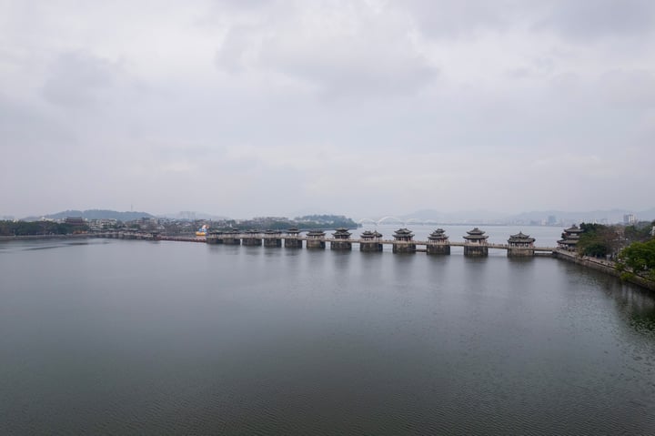 图中是一座横跨湖面的古老桥梁，由多个桥墩组成，每个桥墩上有传统风格的建筑，背景中是城市的建筑和远处的山脉，在多云的天气下显得宁静而美丽。
