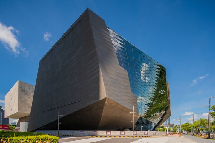 The image features a modern, geometric building with a large, reflective glass facade. The building is situated on a street corner, surrounded by trees and a sidewalk. The sky is clear blue with a few clouds, and the building's design suggests it could be a museum or art gallery. The image does not contain any text or other objects.