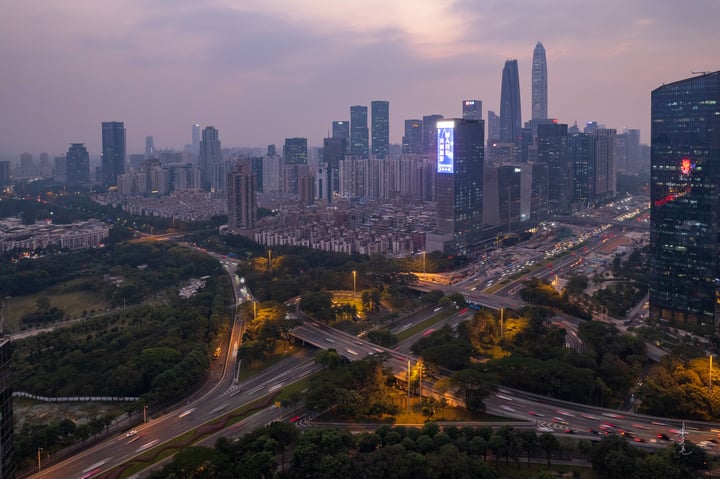 An aerial view of a city at dusk, with tall buildings illuminated by streetlights and a busy highway, surrounded by greenery.