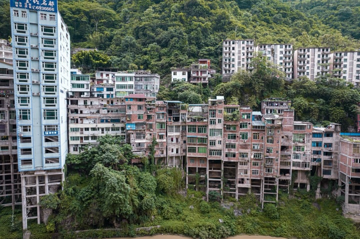 A cityscape on a hillside, with tall buildings of varying heights and colors, surrounded by lush greenery. The river, a brown, choppy body, flows through the city, reflecting the overcast sky.