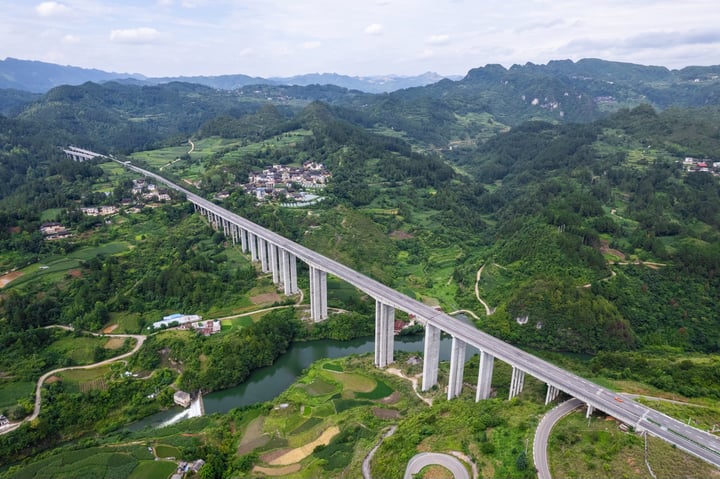 An aerial view of a long concrete bridge spanning a river in a lush green valley, with a town and houses in the distance.