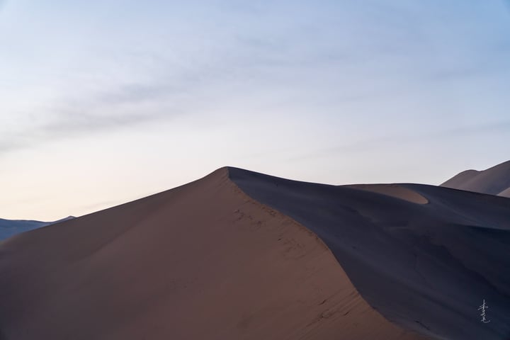 A solitary figure stands on a sandy hill in a vast desert landscape, with a clear blue sky above.
