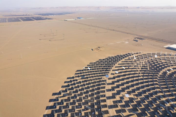 An aerial view of a large solar farm in a desert-like environment, with solar panels arranged in a circular pattern and a large white sun tower in the center. The farm is surrounded by a vast expanse of sandy terrain and a few wind turbines in the distance.