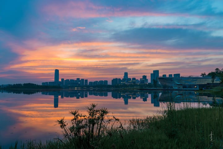 A city skyline at sunset, with tall buildings reflecting on calm water, and a few trees in the foreground.