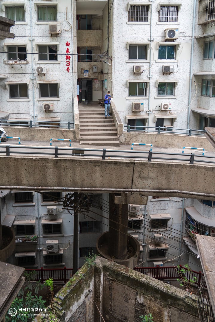 A person in a blue shirt is walking on a concrete staircase in an abandoned apartment building, surrounded by a concrete courtyard with a metal railing and a concrete wall. The building has multiple windows with air conditioning units and a red sign with the letters "S" and "S" on the side. The scene is set against a backdrop of a gray sky.
