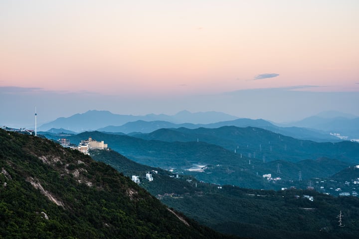 A mountainous landscape with a city on a hill, surrounded by trees and greenery, with a pink and purple sky.