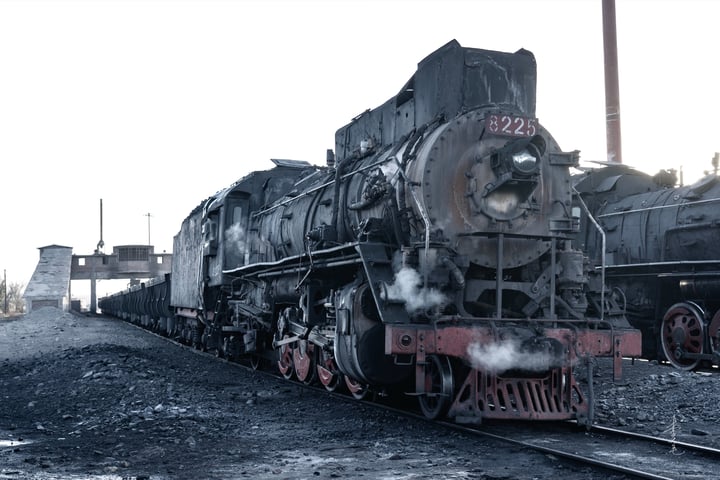 An old black steam locomotive with red wheels is traveling on a railway track, emitting smoke from its chimney.