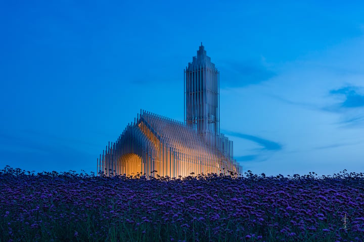 A large white church with a pointed roof and a tall spire stands in a field of purple flowers, illuminated by warm light.