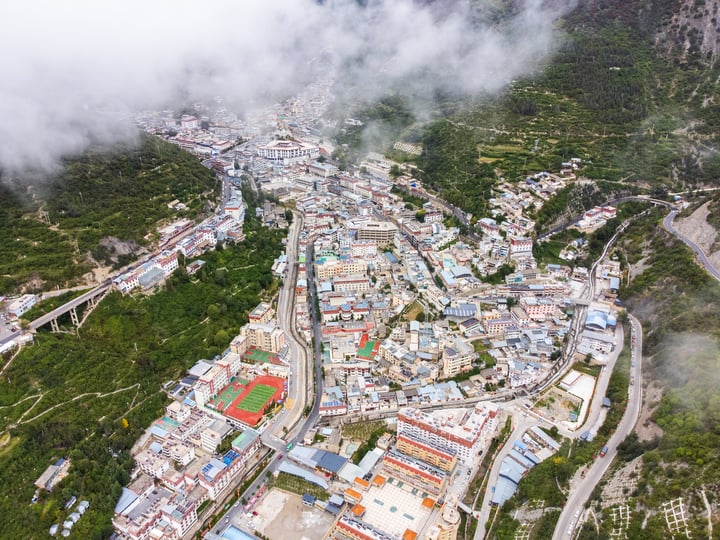 An aerial view of a Deqin County, surrounded by lush greenery and surrounded by mountains. The town is densely populated with houses and buildings, and the roads crisscross the valley. The town is surrounded by trees and hills, creating a serene and picturesque scene.