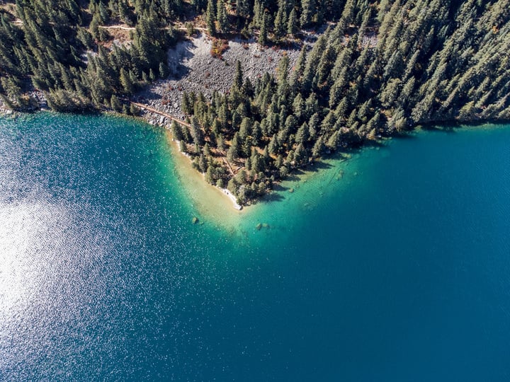 An aerial view of a serene lake surrounded by a dense forest of trees, with a sandy beach in the bottom right corner.