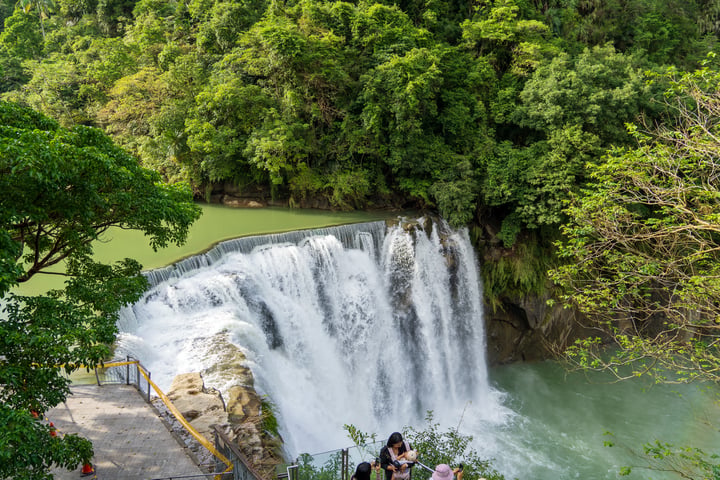 A large waterfall cascades down a cliff, surrounded by lush greenery and a clear blue body of water. People are scattered around the waterfall, some near the edge and others closer to the water. The waterfall is on the right side of the image, with a ramp leading up to it. The scene is serene and picturesque.