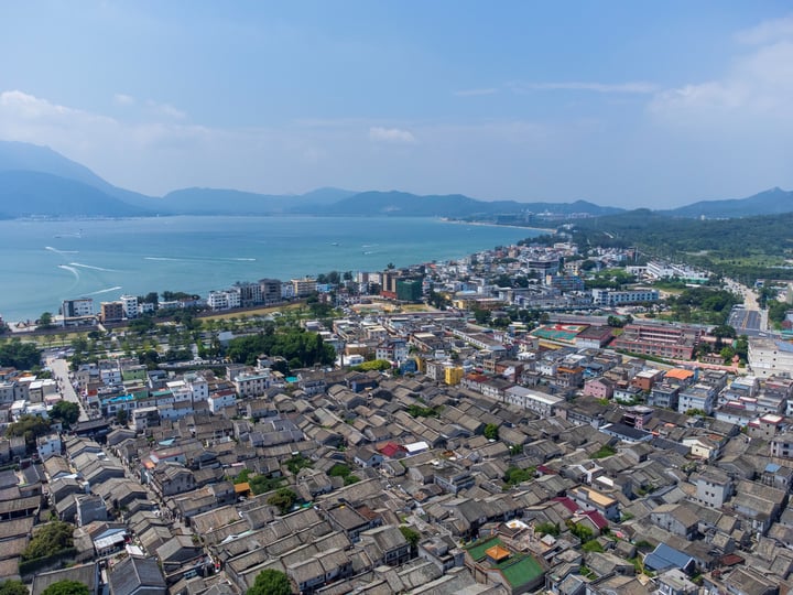 An aerial view of a city with a mix of white and gray buildings, surrounded by mountains and a large body of water.