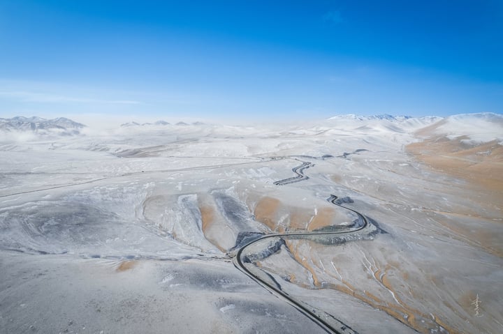 An aerial view of a snowy mountain landscape with a winding road, snow-covered mountains, and a clear blue sky.
