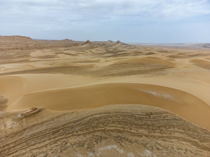 A vast desert landscape with sand dunes, a cloudy sky, and a few small rocks scattered across the landscape.