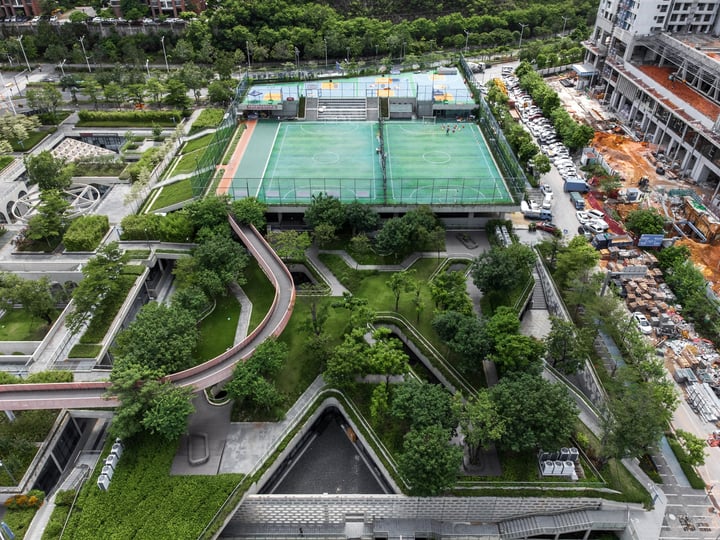 An aerial view of a large green sports facility surrounded by a park, with a tennis court and a basketball court, surrounded by a road and a construction site.