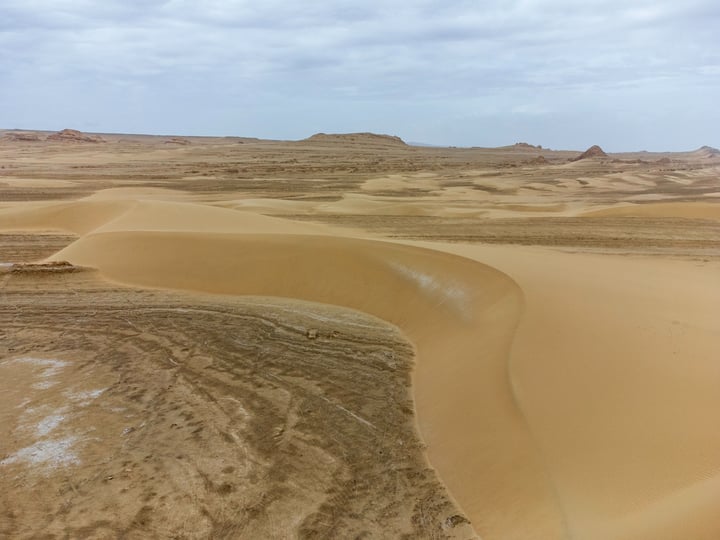 A vast desert landscape with sand dunes, a cloudy sky, and a few small rocks scattered across the landscape.