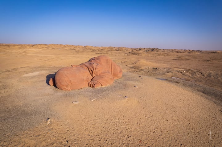 A large, reddish-brown rock lies on the sandy desert, surrounded by small rocks and a clear blue sky.
