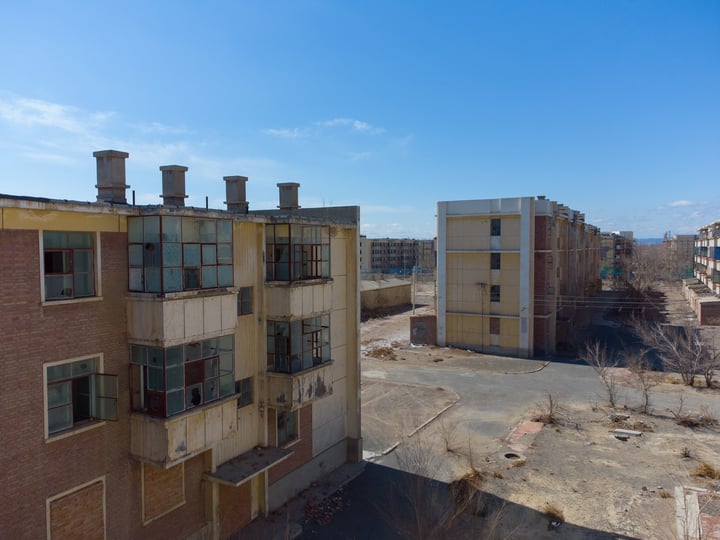 An old, abandoned building with multiple windows and a large chimney on the roof, surrounded by a barren landscape and a clear blue sky.