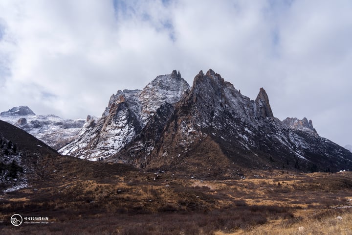 A mountain range with snow-covered peaks and rocky formations, surrounded by a grassy plain and a cloudy sky.