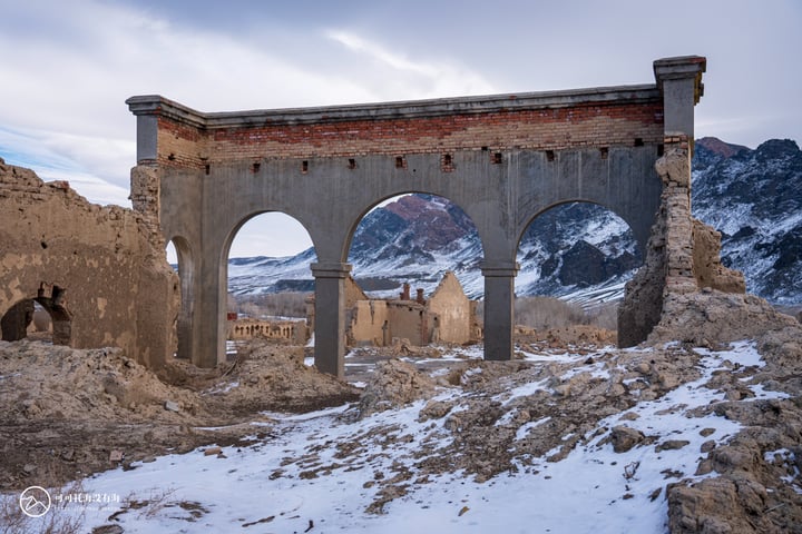An old, weathered stone bridge with three arches stands in a snowy landscape, surrounded by mountains and a cloudy sky.