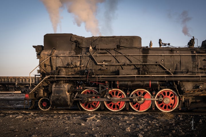 An old black steam locomotive with red wheels is traveling on a railway track, emitting smoke from its chimney.