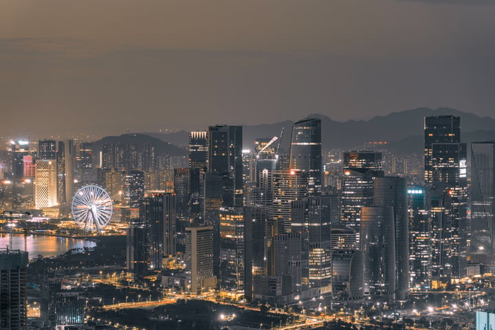 A nighttime cityscape features illuminated skyscrapers, a large Ferris wheel, and mountains in the background.