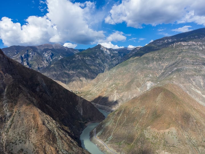 A breathtaking aerial view of a mountainous valley with a winding river, surrounded by majestic mountains and a clear blue sky with fluffy white clouds.