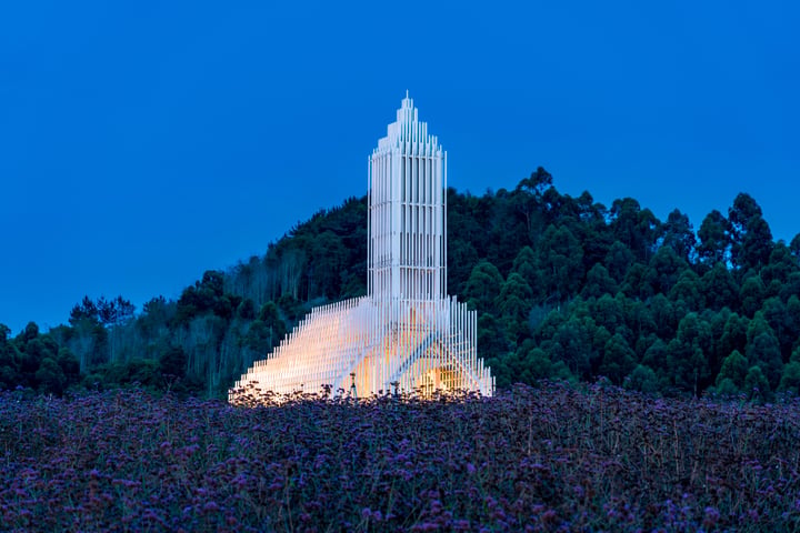 A white, triangular-shaped sculpture stands in a field of purple flowers, illuminated by a warm glow. The sculpture is surrounded by trees, creating a serene and tranquil scene.