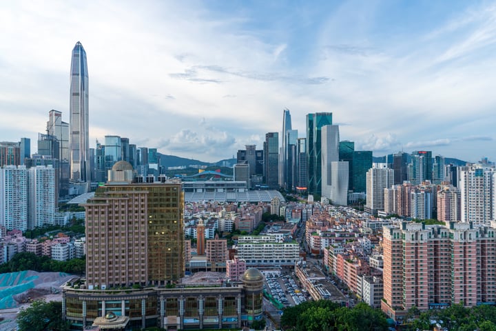 A panoramic view of a bustling city with tall buildings, a skyscraper, and a bridge, surrounded by mountains and a clear blue sky.