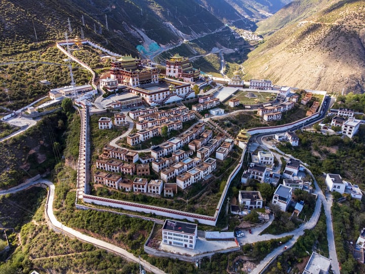 An aerial view of a mountain town with white buildings, red roofs, and a central temple, surrounded by lush greenery and a winding road.