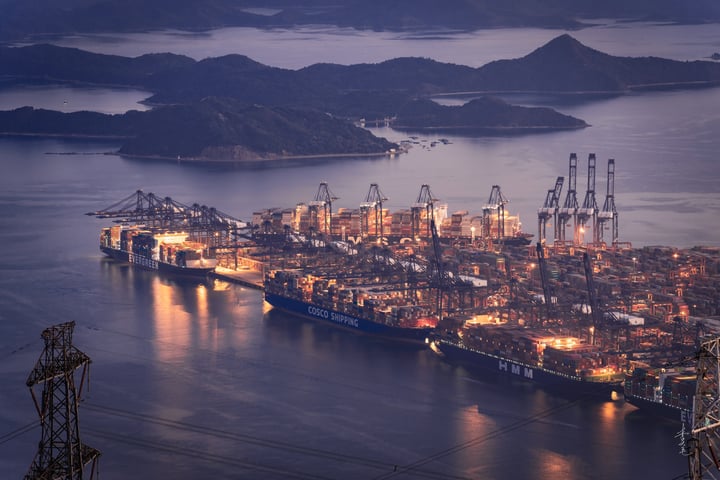 A large cargo ship is docked in a harbor, surrounded by a calm body of water and a distant island with mountains. The ship is filled with cargo and has multiple cranes, indicating it's a large ship. The sky is dark blue, and the ship is illuminated by lights.