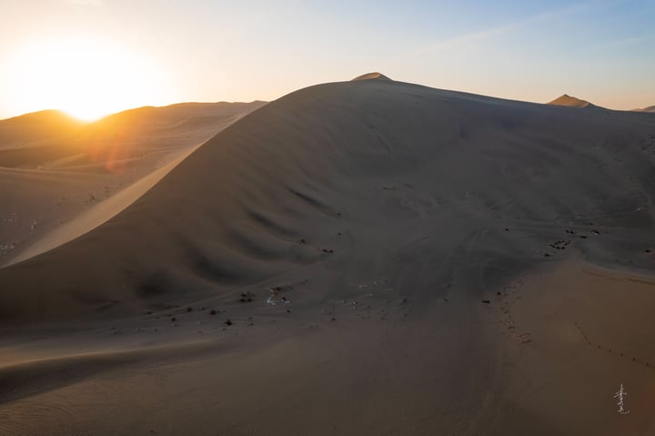 A serene desert landscape at sunset, with a large sand dune in the foreground and smaller dunes in the background. The sun is setting, casting a warm glow on the sand and creating a tranquil atmosphere.