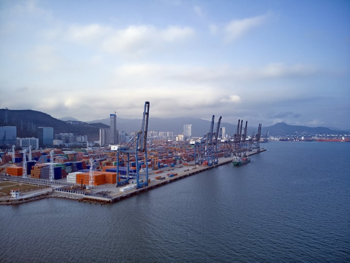 An aerial view of a bustling port with colorful shipping containers, cranes, and a city skyline in the background.