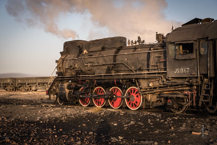 An old black steam locomotive with red wheels is traveling on a railway track, emitting smoke from its chimney.