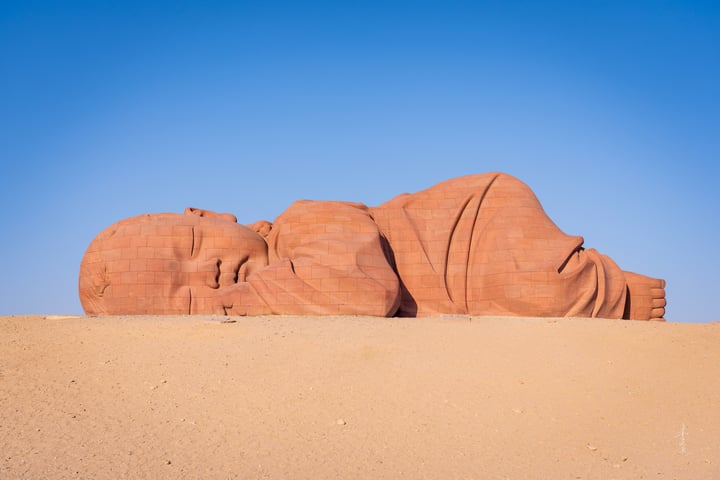 A large, red, weathered sculpture of a person lying on their back in a desert-like environment, with a clear blue sky in the background.