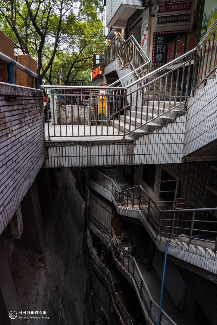 A narrow alleyway in a city, with a metal staircase leading to a building on the right side, surrounded by trees and people walking around.