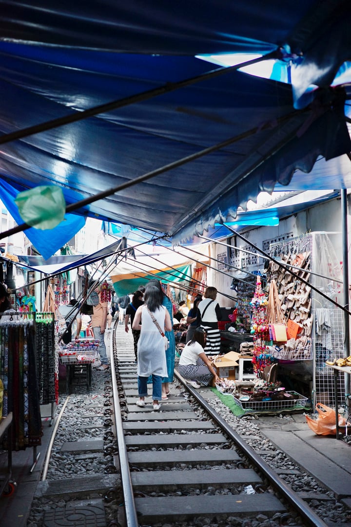 A bustling market in Thailand features a variety of stalls under a blue tarp, with people browsing and shopping.