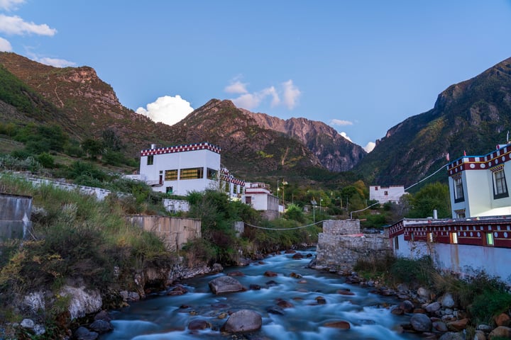 A serene mountain landscape with a river flowing through it, surrounded by white buildings with red and white accents, and a clear blue sky with white clouds.