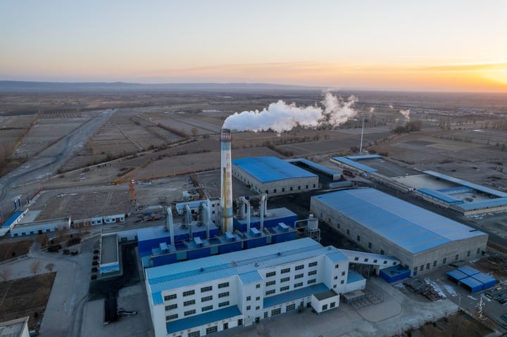 An aerial view of a large industrial complex with multiple buildings, including a large factory with smokestacks and a large factory with a large factory. The complex is surrounded by a large city with buildings of varying heights and colors. The sky is clear with a few clouds.