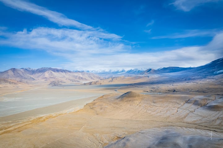 A vast, sandy desert landscape with a river in the foreground, mountains in the background, and a blue sky with white clouds.
