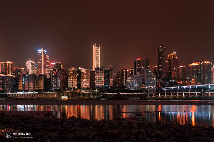 A nighttime cityscape is reflected in calm water, with tall buildings illuminated by lights and a bridge crossing the river.