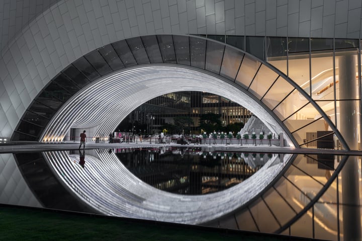 A modern building with a curved white metal archway and reflective pool in front, illuminated by white lights, is surrounded by a dark gray building and a solitary figure walking on the sidewalk.