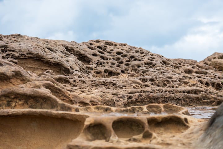 A rocky landscape with a cloudy sky, a large rock with holes and cracks, and a small body of water in the background.
