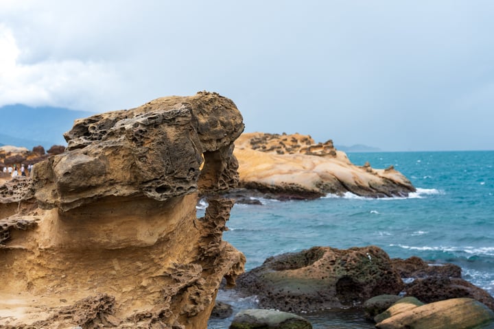 A rocky coastline with a large, weathered rock formation, a small cave, and a mountain range in the background.