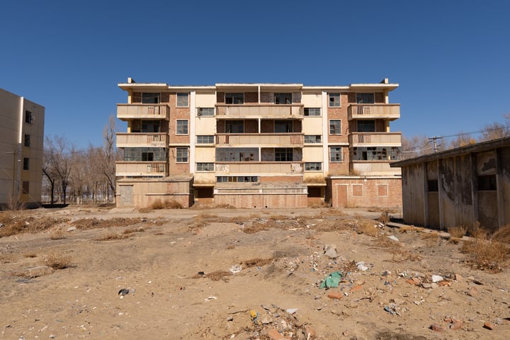 An old, dilapidated apartment building with numerous windows and balconies stands in a barren, dusty area surrounded by a dirt field and bare trees.