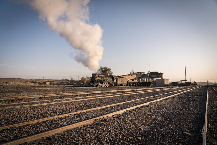 A black train engine is on a set of train tracks, with a small building and a smokestack on the left side, and a clear blue sky above.